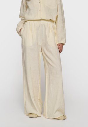 Mujer vistiendo pantalones sueltos de pierna ancha color crema con cintura elástica, combinados con una blusa a juego de manga larga y botones, y zapatos de punta cuadrada.