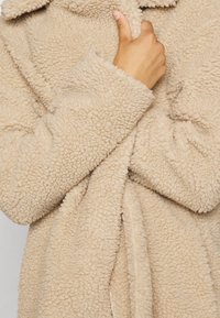 Hellbeiger Fleece-Mantel mit flauschiger Textur, langen Ärmeln und Schalkragen. Der Stoff ist weich und hat ein gemütliches Aussehen.