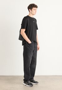 Black t-shirt with a small white logo, dark grey jeans, and black sneakers. Model holds a black tote bag; casual fit with relaxed shapes.