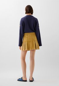 Person, der mit dem Rücken steht und ein navyfarbenes Langarmshirt, einen senfgelben plissierten Rock und blaue Slipper trägt.