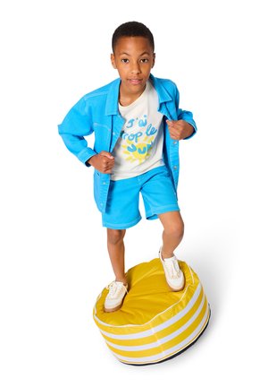 Garçon portant une veste bleue et un short, debout sur un coussin rond rayé jaune et blanc, tenant la veste ouverte, souriant légèrement.