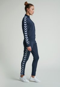 Marinblå sporttracksuit med full-zip jacka med ståkrage och sidosträck i chevron-mönster; tillsammans med matchande tighta byxor.