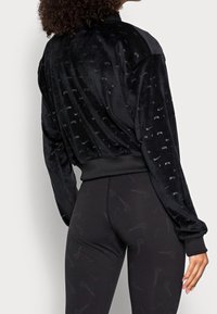 Kvinna som bär en svart velourjacka och svarta leggings med subtila Nike-swoosh-mönster, visad bakifrån.