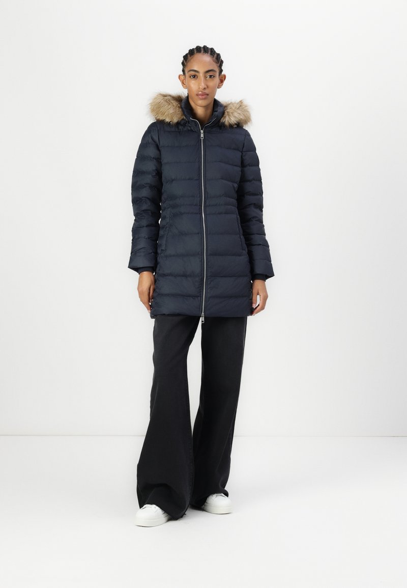 Marineblauer Puffer-Mantel mit Kapuze aus Kunstpelz, Reißverschluss vorne und gerippten Bündchen. Kombiniert mit schwarzen weit geschnittenen Hosen und weißen Sneakern. Schlichtes Design.
