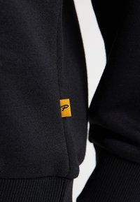 Sweatshirt noir avec des poignets et un ourlet côtelés, présentant une petite étiquette jaune avec un logo sur le côté. Texture douce et détails de design simples.