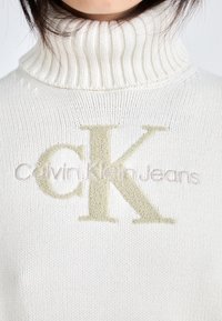 Weißer, gerippter Pullover mit hohem Rollkragen, versehen mit einem auffälligen beigen "CK"-Logo und kleinerem Schriftzug "Calvin Klein Jeans" darunter.