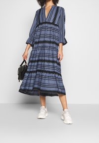 Robe longue noire à motifs géométriques bleus, col en V et manches évasées. Associée à des baskets blanches et un petit sac à main noir.