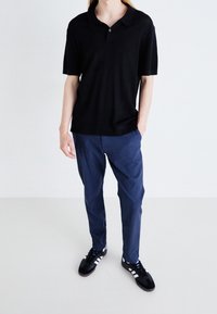 Černé polo tričko s krátkým rukávem a ribbovanou texturou, v kombinaci s tmavě modrými kalhotami. Model má na sobě černé tenisky s bílými pruhy.