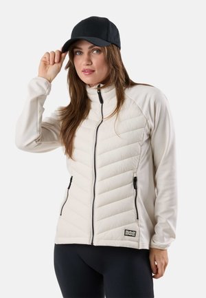 Kobieta w jasnobeżowej pikowanej kurtce z czarnymi zamkami błyskawicznymi, czarnych legginsach i czarnej czapce z daszkiem, poprawiająca czapkę jedną ręką.