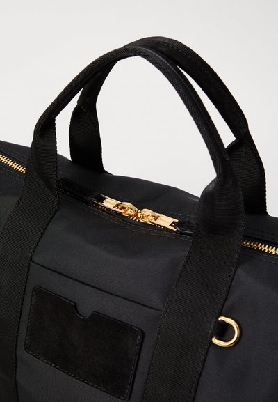 Black fabric bag with dual handles, gold zipper hardware, and a suede patch detail. Features a structured design and smooth texture.