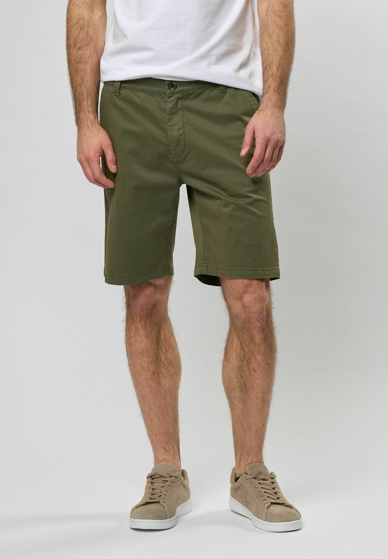 Pantalones cortos de algodón color verde oliva con bolsillos laterales, una cremallera y un cierre de botón. Combinados con una camiseta blanca y zapatillas beiges.