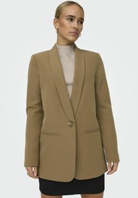 Hnědý blazer se šálovým límcem, s jedním knoflíkovým zapínáním, dvěma předními kapsami a výraznými ramenními liniemi. Nesený přes ribbovaný béžový top.