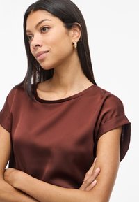 Top in satin marrone con una texture liscia, scollo rotondo e maniche corte arrotolate, che presenta una silhouette rilassata e una morbida caduta.