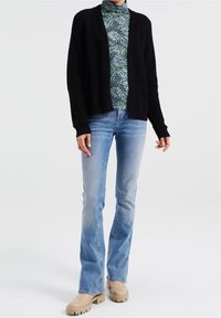 Svart stickad kofta med öppen framsida, bärs över en mönstrad, grön och blå polotröja. Ljusblå utsvängda jeans och beigea klumpiga skor.