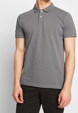 Homme portant un polo gris ajusté avec deux boutons et un pantalon foncé, debout devant un fond clair uni.