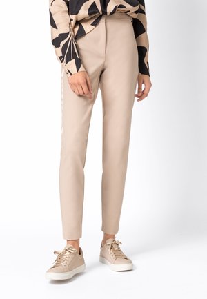Personne portant un pantalon beige à jambes droites, des baskets beige assorties et une chemise à manches longues à motif abstrait noir et beige.