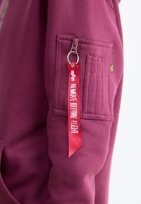 Manga de chaqueta burdeos con un bolsillo con cremallera y una etiqueta de tela roja de llavero que dice "RETIRAR ANTES DEL VUELO" unida al tirador de la cremallera.