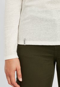 Weißes Langarm-T-Shirt mit strukturierter Oberfläche, ausgestattet mit einem kleinen Markenetikett am Saum, kombiniert mit dunkelgrünen Hosen.