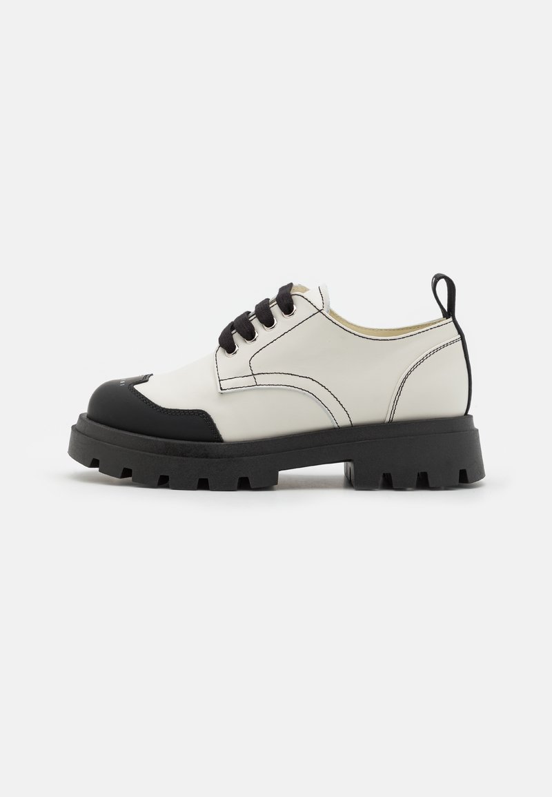Chaussure basse blanche avec embout noir, semelle noire épaisse, lacets noirs, surpiqûres contrastantes et languette à l’arrière.