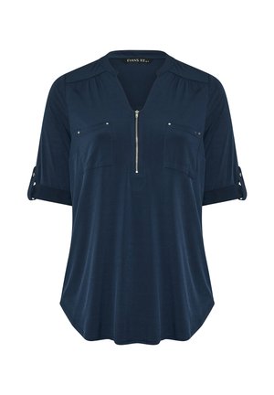 Tunique bleu marine avec des manches courtes retroussées, fermeture éclair à l'avant, deux poches poitrine et de subtils plis aux épaules. Texture du tissu douce.