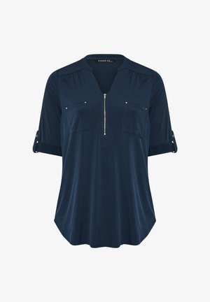 Granatowa tunika z krótkimi, podwiniętymi rękawami, z zamkiem błyskawicznym z przodu, dwiema kieszeniami na piersi i subtelnymi plisami na ramionach. Miękka tekstura materiału.