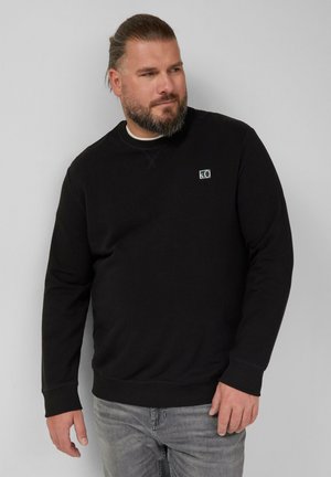 Schwarzer Pullover mit rundem Halsausschnitt und gerippten Bündchen, kombiniert mit hellgrauen Jeans. Zeigt ein kleines Logo auf der Brust.