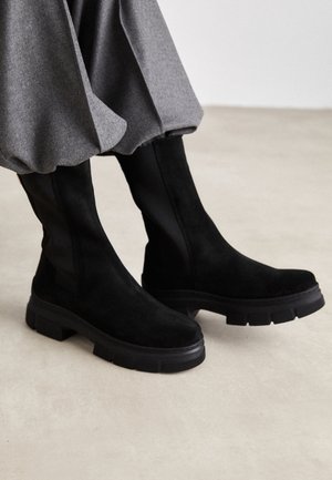 Bottes cheville en daim noir avec semelles épaisses portées avec un pantalon gris à revers sur un fond de sol clair.