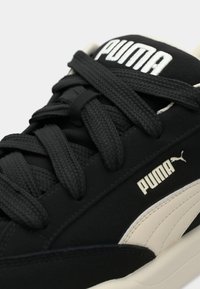 Black sneaker featuring a matte texture, thick black laces, and a white Puma logo. The design includes a cream-colored accent panel.