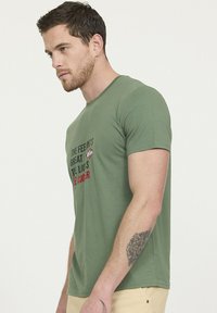 T-shirt en coton vert olive à manches courtes, avec du texte en gras rouge et noir sur la poitrine. Encolure ras du cou classique et texture douce.