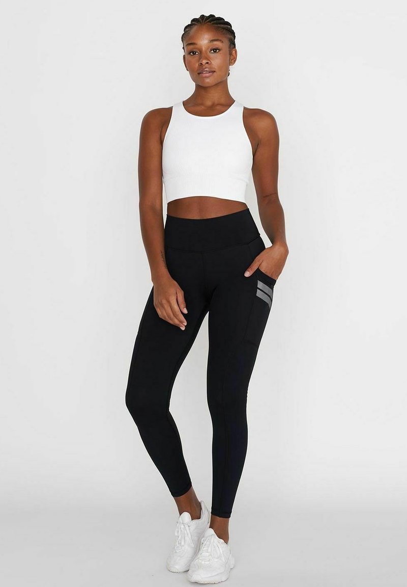 Leggings noires taille haute avec poches latérales et rayures grises, associés à un crop top blanc à côtes. Des chaussures de sport blanches complètent le look.