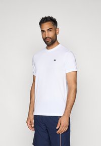 T-shirt bianca a girocollo in tessuto leggero, con un piccolo logo ricamato verde sul petto. Maniche corte, design minimale.