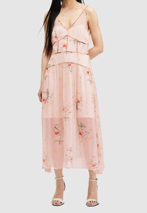 Frau trägt ein ärmelloses, rosafarbenes Midikleid mit floralen Mustern, dünnen Trägern und weißen Sandalen mit Absatz vor einem schlichten Hintergrund.