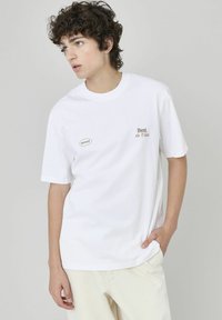 T-shirt en coton blanc avec le texte "Meilleur de sa catégorie" en marron et une étiquette de marque. Manches courtes et coupe décontractée, associée à un pantalon de couleur claire.