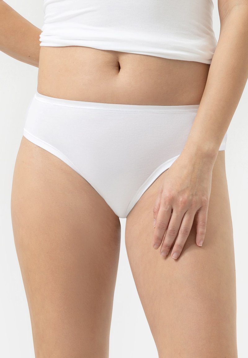Femme portant une sous-vêtements en coton blanc uni et un haut blanc, avec une main reposant sur sa cuisse contre un fond blanc.