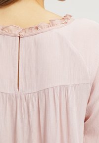 Dos d'un chemisier rose clair avec une petite fermeture à bouton, un col à volants et un tissu froncé sous la couture de l'empiècement.