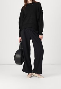 Schwarzer übergroßer Pullover, weite schwarze Hose und beigende flache Schuhe. Hält eine runde schwarze Handtasche. Minimalistisches Design und strukturierter Stoff.