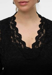Top en dentelle noire avec un col en forme de vagues, des motifs floraux complexes et une finition texturée. Le tissu est transparent avec des détails en couches.