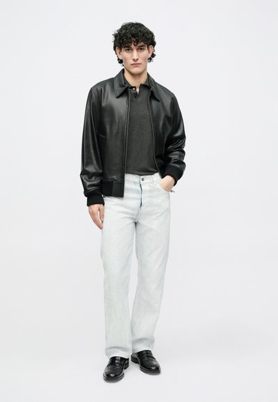Μαύρο δερμάτινο μπουφάν bomber πάνω από σκούρο γκρι πόλο, σε συνδυασμό με τζιν ελαφριάς πλύσης και ίσιας γραμμής και μαύρα μοκασίνια. Ομαίες υφές, ελάχιστες λεπτομέρειες.