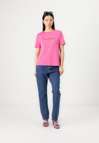 Rosa bomullströja med broderad logotext framtill, som matchas med blå raka jeans och lila sandaler med remmar.