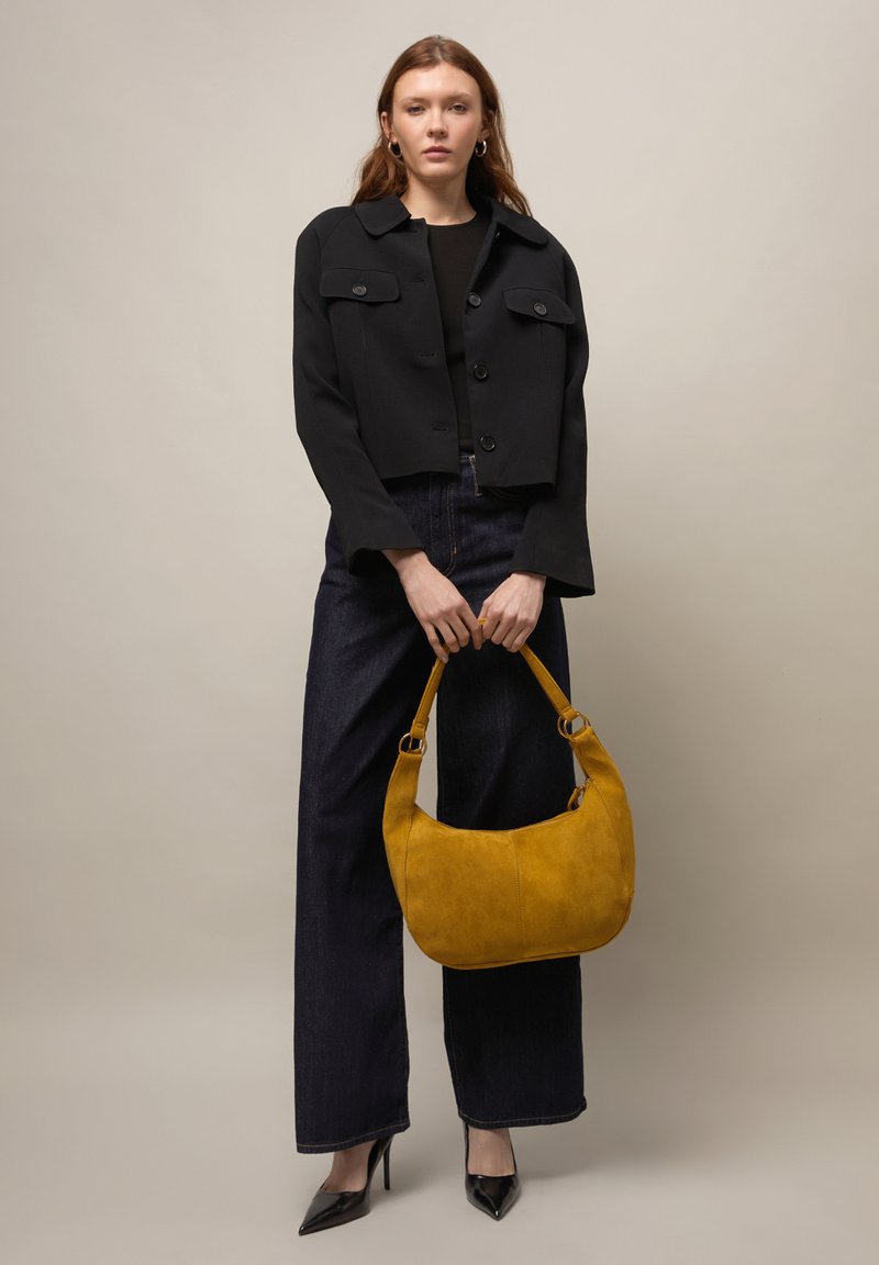 Donna con giacca nera corta e jeans a gamba larga, che tiene una borsa giallo senape, in piedi contro uno sfondo uniforme.