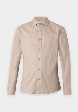 Homme portant une chemise beige à manches longues boutonnée, un pantalon marron et des chaussures noires, debout avec une main dans le dos.