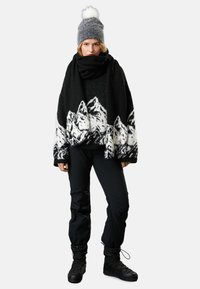 Mustan ja valkoisen villahuovan poncho, jossa on vuorimaisema-kuvio, yhdistetty mustaan huiviin ja istuviin mustiin housuihin. Viimeistelty mustilla, eristetyillä nauhakenkillä.