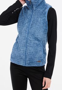 Blauer flauschiger Weste mit hohem Kragen, durchgehendem Reißverschluss vorne und Seitentaschen. Weiche Textur; kontrastierende blaue Einfassung entlang der Armausschnitte. Schwarzes Langarmshirt darunter.