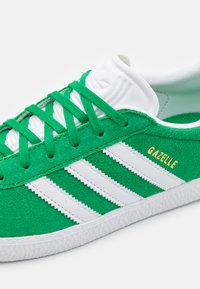 Green suede sneaker with three white stripes, textured surface, and gold "Gazelle" logo; white rubber sole and laces complete the design.