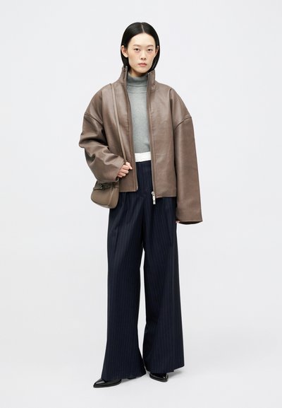 Νεαρή γυναίκα με καφέ oversized δερμάτινο μπουφάν, γκρι ζιβάγκο, ναυ blue ριγέ παντελόνι φαρδιάς γραμμής, μαύρα κομψά παπούτσια και κρατώντας μια καφέ τσάντα ώμου.