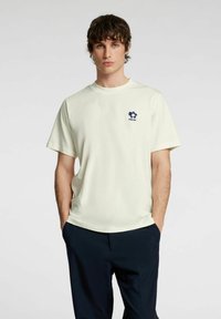 Lysegul T-shirt med korte ærmer, rund halsudskæring og et mørkeblåt blomstermotiv på venstre bryst; stoffet ser blødt og glat ud.