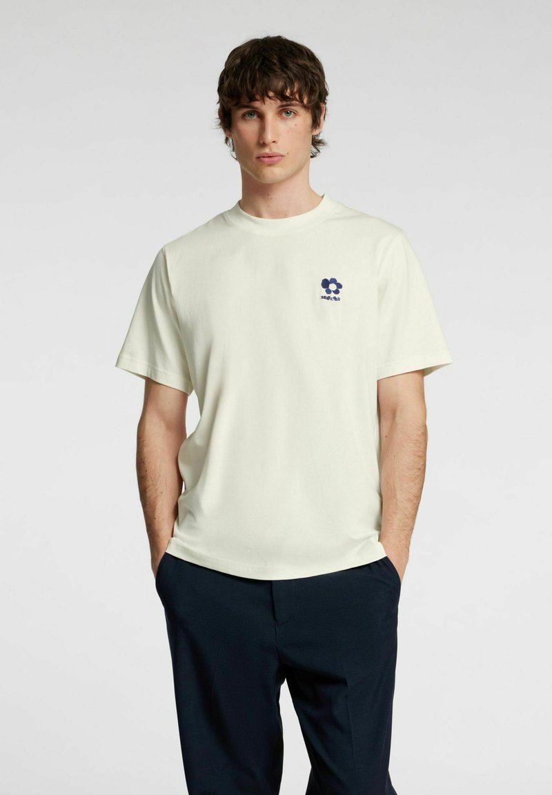 Lysegul T-shirt med korte ærmer, rund halsudskæring og et mørkeblåt blomstermotiv på venstre bryst; stoffet ser blødt og glat ud.