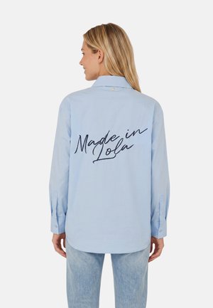 Femme aux cheveux blonds portant une chemise bleu clair avec l'inscription "Made in Lola" dans le dos, associée à un jean bleu clair, de dos.