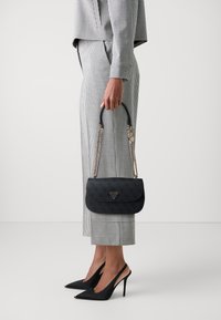 Borsa nera strutturata con forma curvata, tracolla in catena dorata e accentuazione del logo. Indossata con pantaloni a scacchi grigi e tacchi neri.