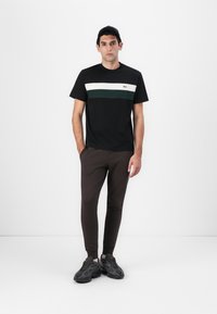 Schwarzes T-Shirt mit grünen und weißen horizontalen Streifen, kombiniert mit braunen, schmal zulaufenden Jogginghosen und dunklen Sneakers. Einfaches, lässiges Design.
